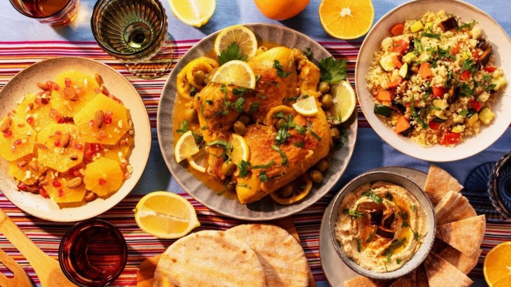 Overhead view of a bright spread: lemon-garnished roast chicken, orange segments with pomegranate, quinoa salad, hummus with pita, and lemon slices on a striped tablecloth.