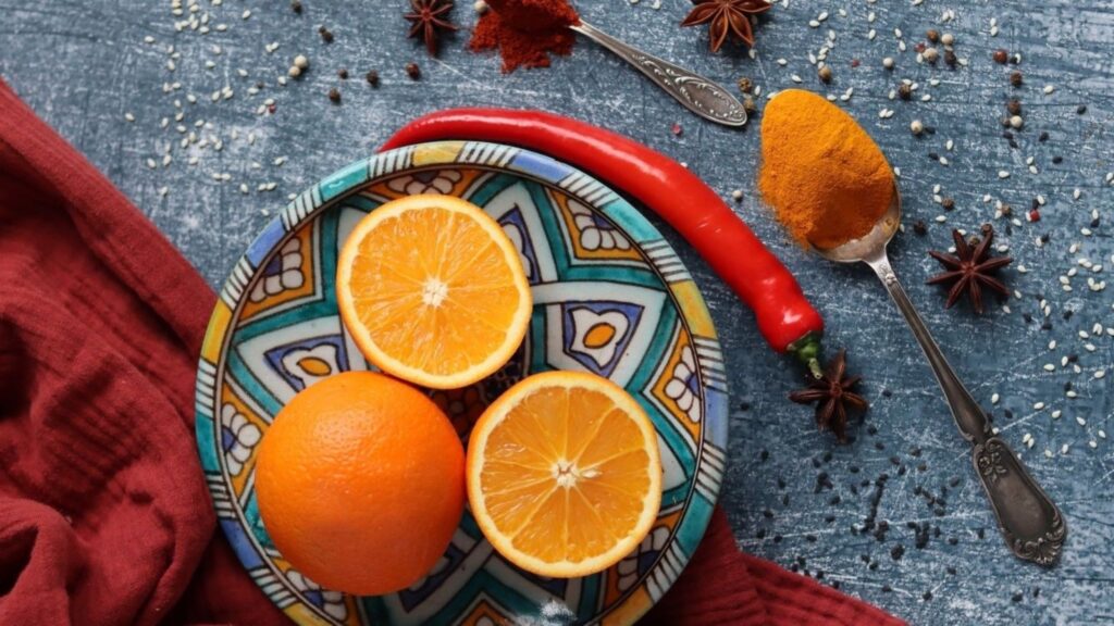 Plate with sliced oranges and a whole orange, a red chili pepper, and spices on a blue rustic surface. Decorative, colorful culinary scene.