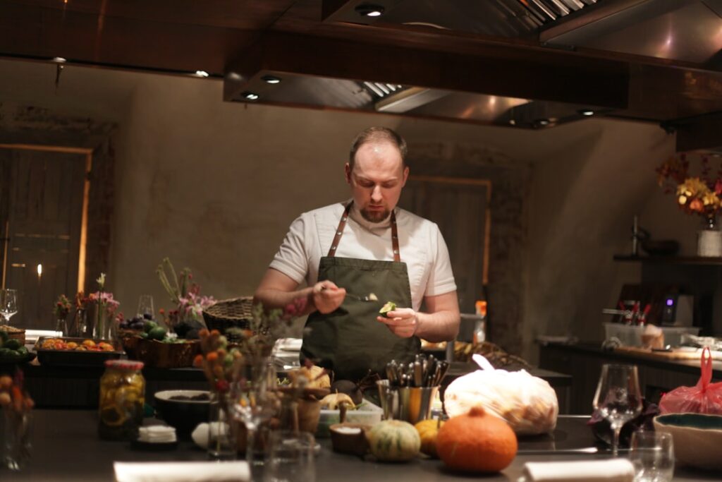 Chef in a white shirt and green apron plates food at a busy kitchen counter, surrounded by vegetables and bowls.