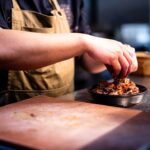 A chef carefully assembles a balanced grain bowl with layered grains, vegetables, and fresh herbs on a wooden counter