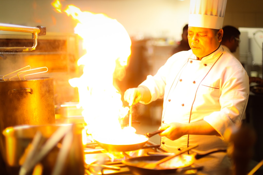 A chef holds tongs over an open flame while charring summer peppers on a gas stovetop in a professional kitchen.