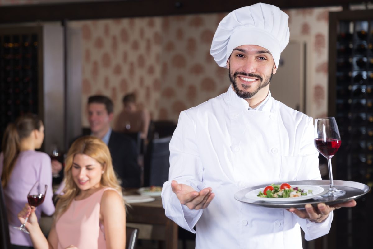A chef steps out of a small restaurant kitchen to welcome loyal regulars at their table
