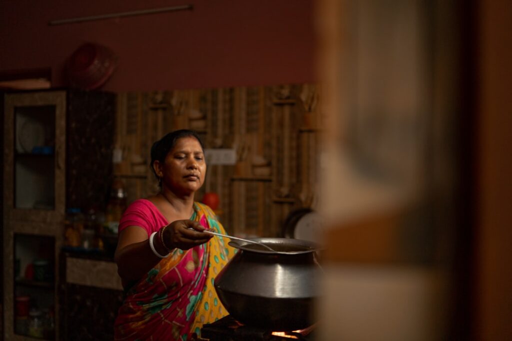 Indian woman in a colorful sari cooking over a metal pot on a stove, reaching with a ladle in a dimly lit kitchen.