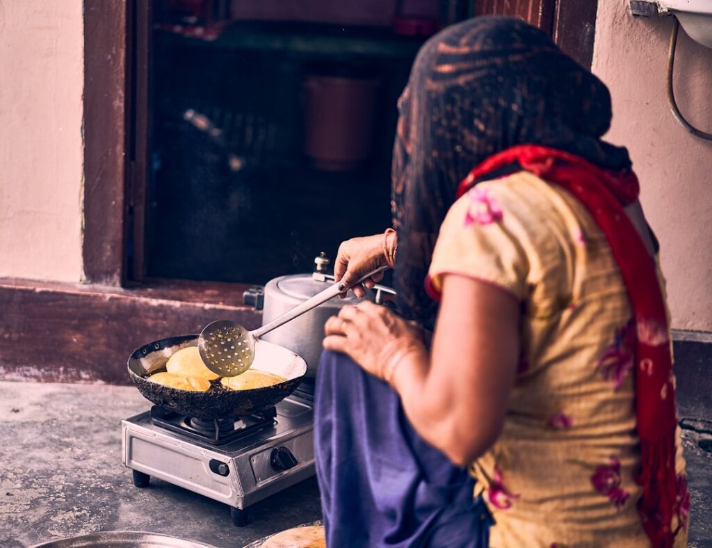 Woman in a colorful dress and red scarf frying round flatbreads on a pan over a portable gas stove outside a doorway.