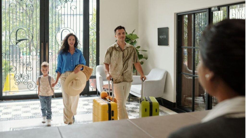 Family of three walks into a bright hotel lobby with yellow and green rolling suitcases as they enter through ornate glass doors.