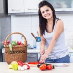 A woman carefully arranges containers of picnic recipes into a wicker basket on a kitchen, ready for a June outdoor outing.