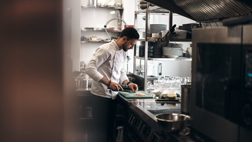 Chef in a white jacket chopping ingredients on a green cutting board at a stainless steel prep counter in a busy kitchen