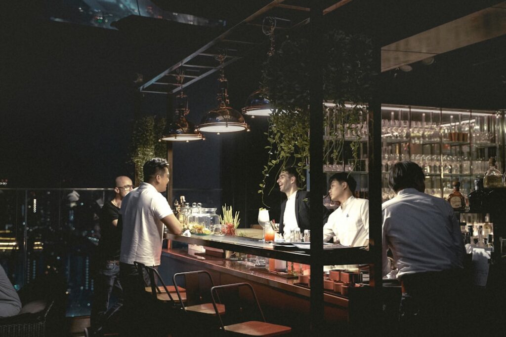 Nighttime rooftop bar with a bartender and a customer leaning on the illuminated counter, city lights in the background.