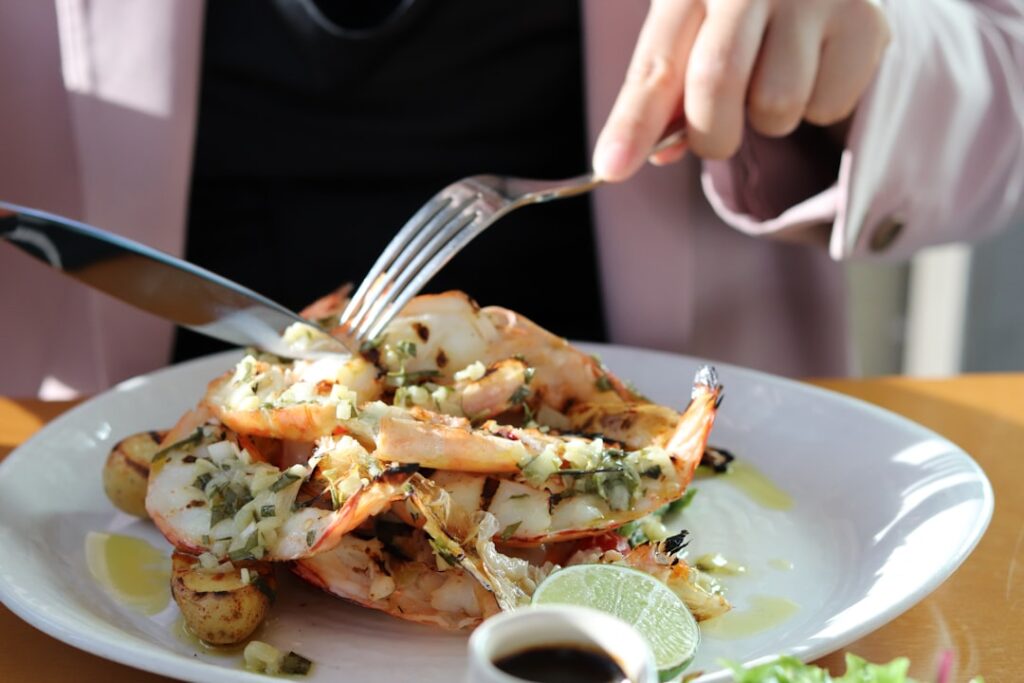 Plate of shrimp topped with fresh herbs and a lime wedge, being cut with a fork and knife at a wooden table.