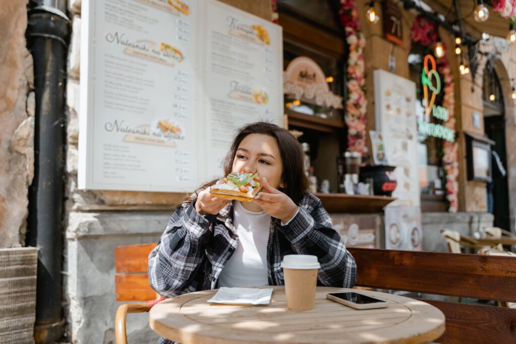 Woman in a plaid shirt eating a sandwich at an outdoor cafe; coffee cup and smartphone on the table with menus behind her.
