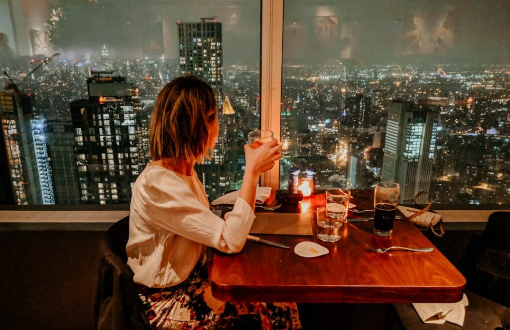 Woman seated at a candlelit restaurant table, toasting with a glass as city lights glow through a window behind her at night.
