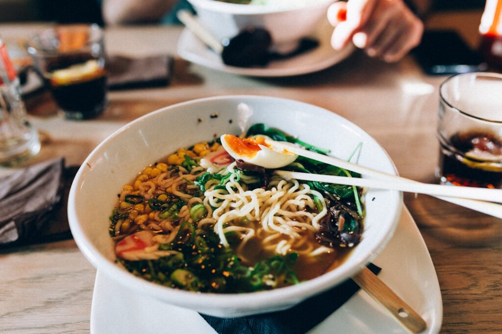 Bowl of ramen with noodles, corn, greens, and a half-boiled egg, chopsticks resting on top on a wooden table in a dining setting.