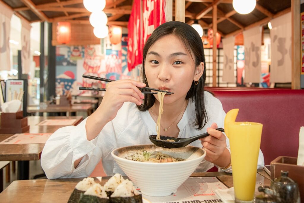 Young woman eats ramen in a casual restaurant, lifting noodles with chopsticks over a large bowl of noodles on the table.