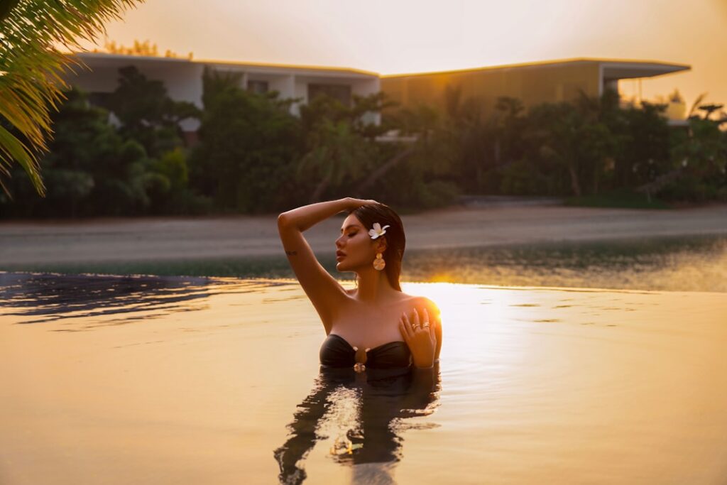 Woman in a black bikini standing chest-deep in a sunset-lit pool, hand resting on her head with a flower in her hair.