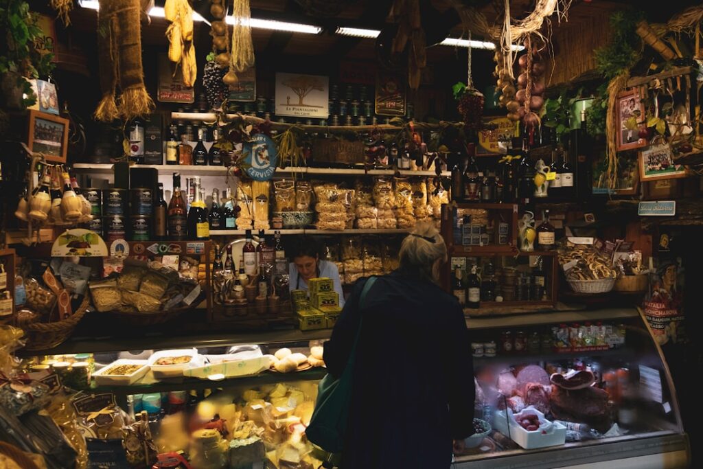 Cozy market stall with shelves of bottles, jars, and dried goods; a customer browses the counter display behind glass.