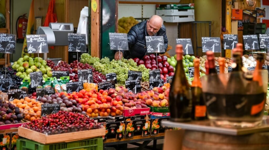 Colorful fruit stall at a market with a worker behind the display and chalkboard price signs visible among the produce.