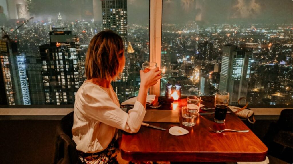 Woman with short hair in a white blouse sits at a wooden table by a window, holding a drink, overlooking a city skyline at night with lit buildings and candles on the table.