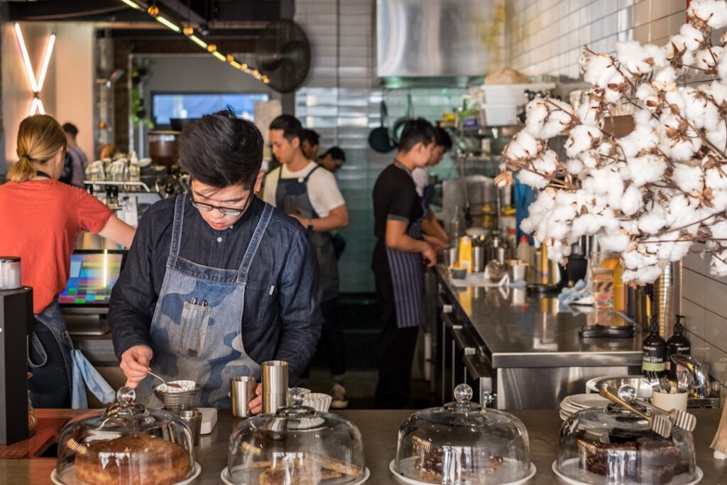 Barista in a denim apron scooping dessert at a busy cafe counter with glass cake domes in the foreground.