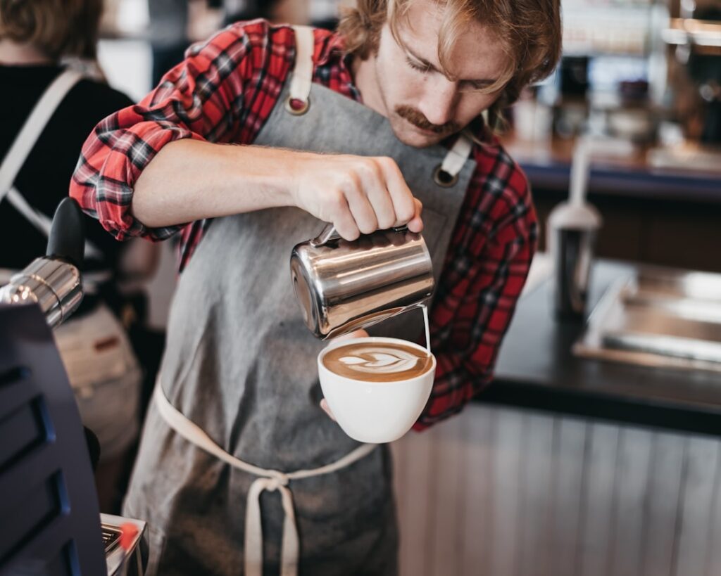 Barista in a plaid shirt pours steamed milk from a metal pitcher into a latte in a white cup at a cafe