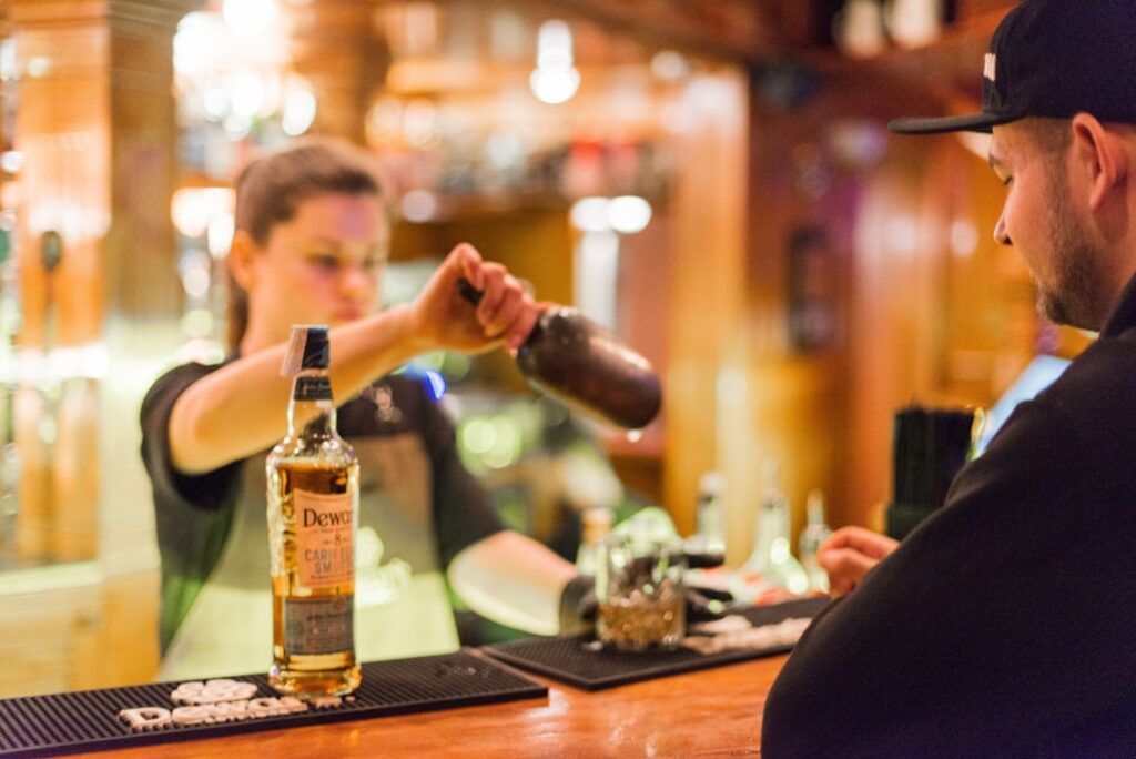 Bartender pouring whiskey from a bottle into a shaker for a customer at a bar by the counter.