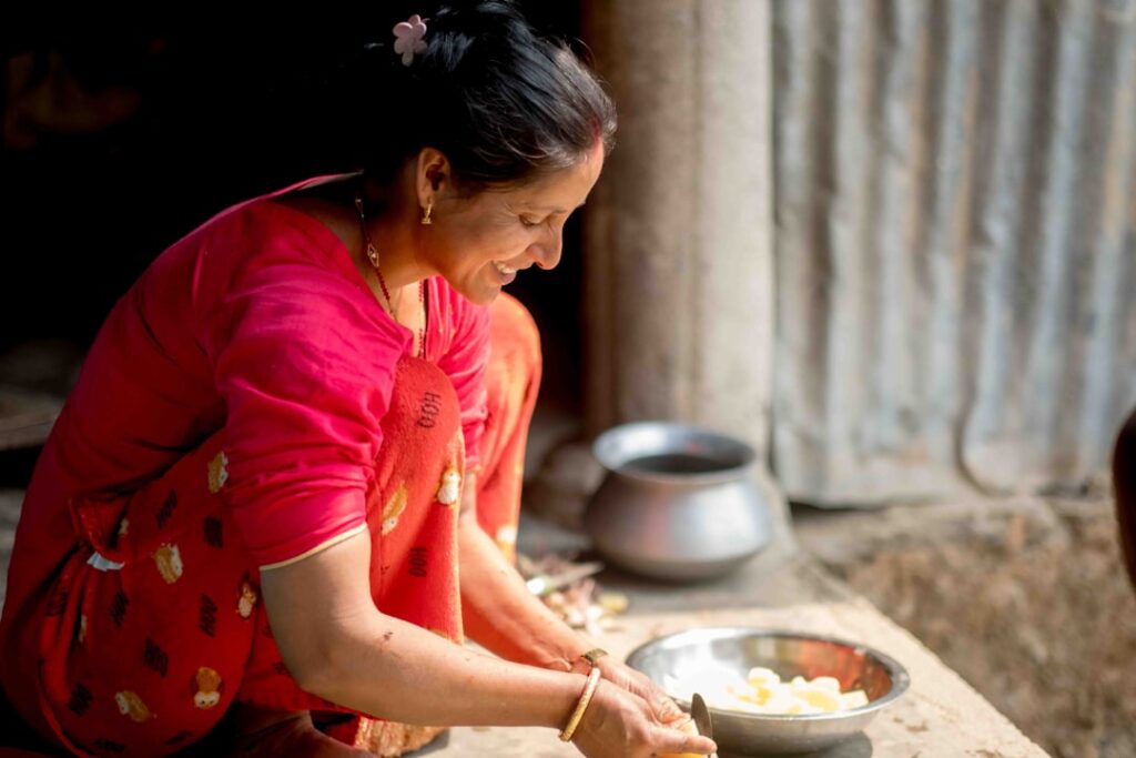 Woman in a bright pink saree chops vegetables in a rustic outdoor kitchen, with metal bowls nearby.
