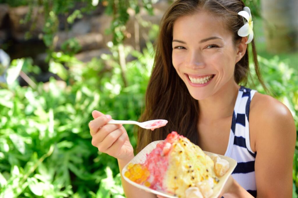 Smiling woman outdoors with a white flower in her hair holding a bowl of colorful shaved ice dessert on a spoon.