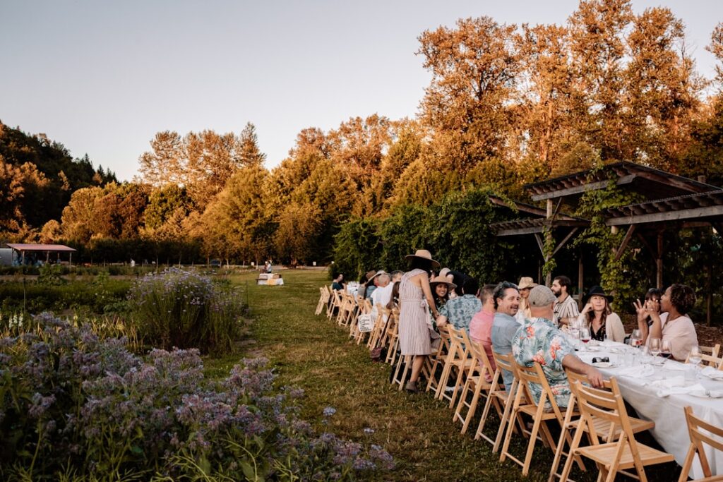 Long outdoor garden party or wedding reception with a long table of guests dining under a wooden pergola as autumn trees glow in the background