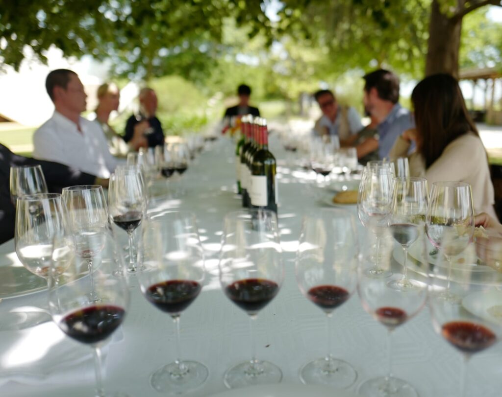 Outdoor al fresco lunch with a long table, wine glasses, and bottles as guests chat under trees.