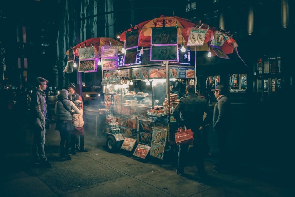 Nighttime street food cart with illuminated signs and steam rising as customers queue nearby on a city sidewalk.