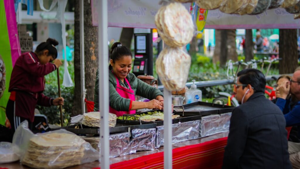 Street food vendor flipping food on griddles at a busy outdoor cart, with customers seated nearby under colorful awnings.