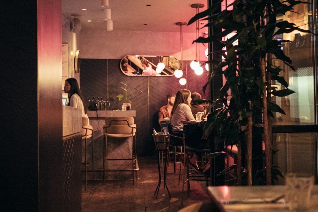 Café interior at night with warm pink lighting, patrons seated at tables near a bar; leafy plants in the foreground.