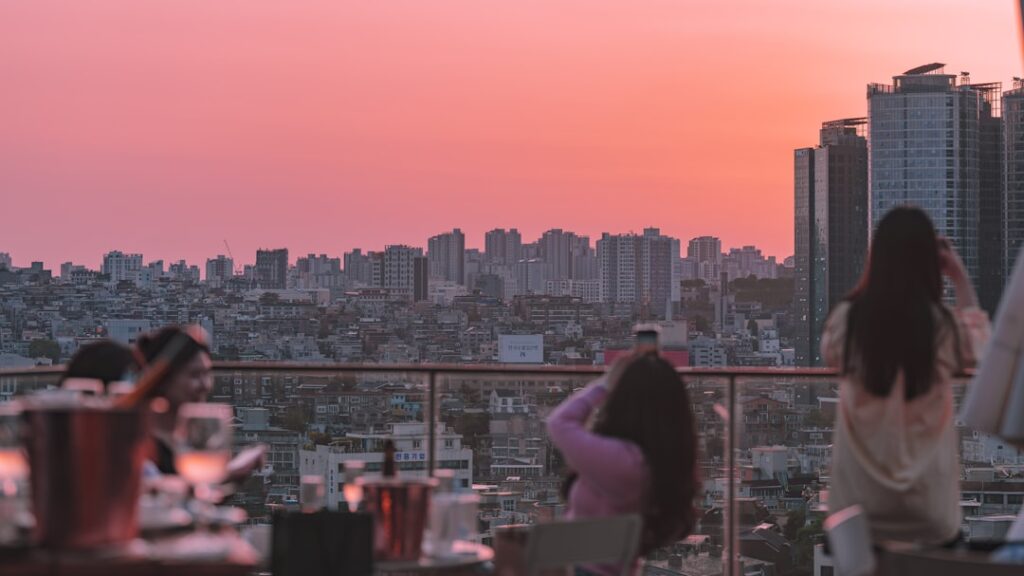 People on a rooftop terrace at sunset, overlooking a dense city skyline with a pink-orange sky.