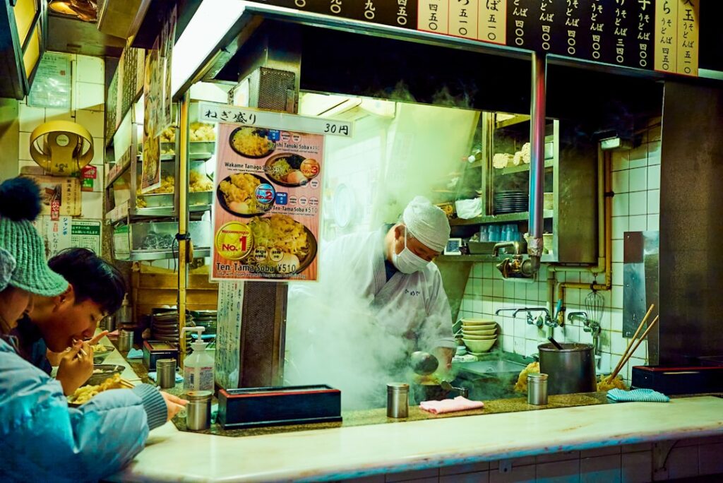 Open Japanese noodle shop counter with a chef in a white hat and mask cooking amid rising steam as customers eat at the bar to the left.
