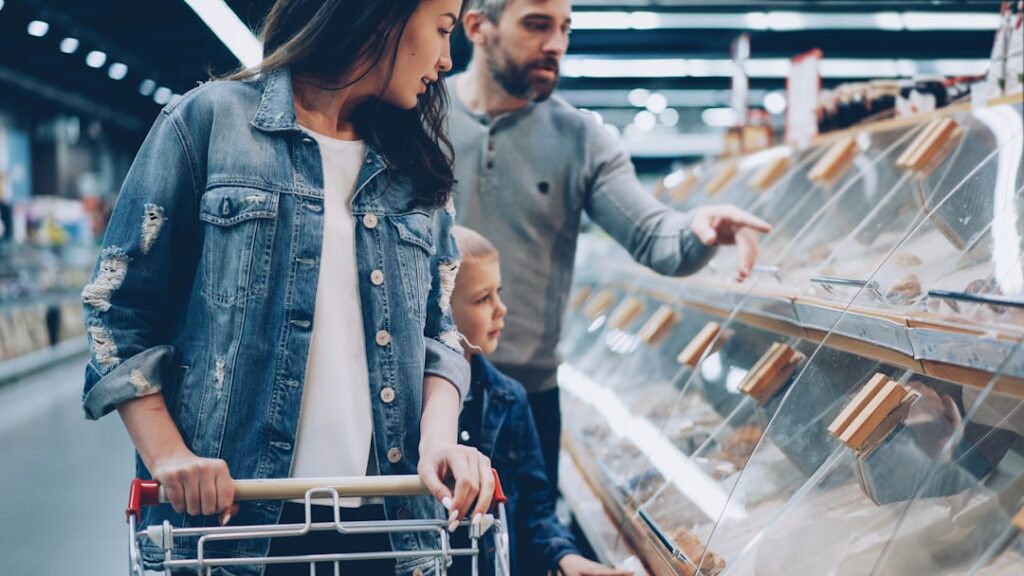 Mother pushes a shopping cart with her young son nearby as the father points to pastries in a glass bakery display case in a store.