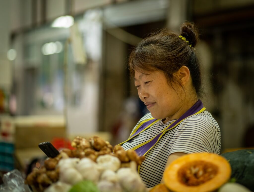 Market vendor, a woman in a striped shirt looking at her phone while standing near piles of produce including garlic and ginger in a busy market stall.