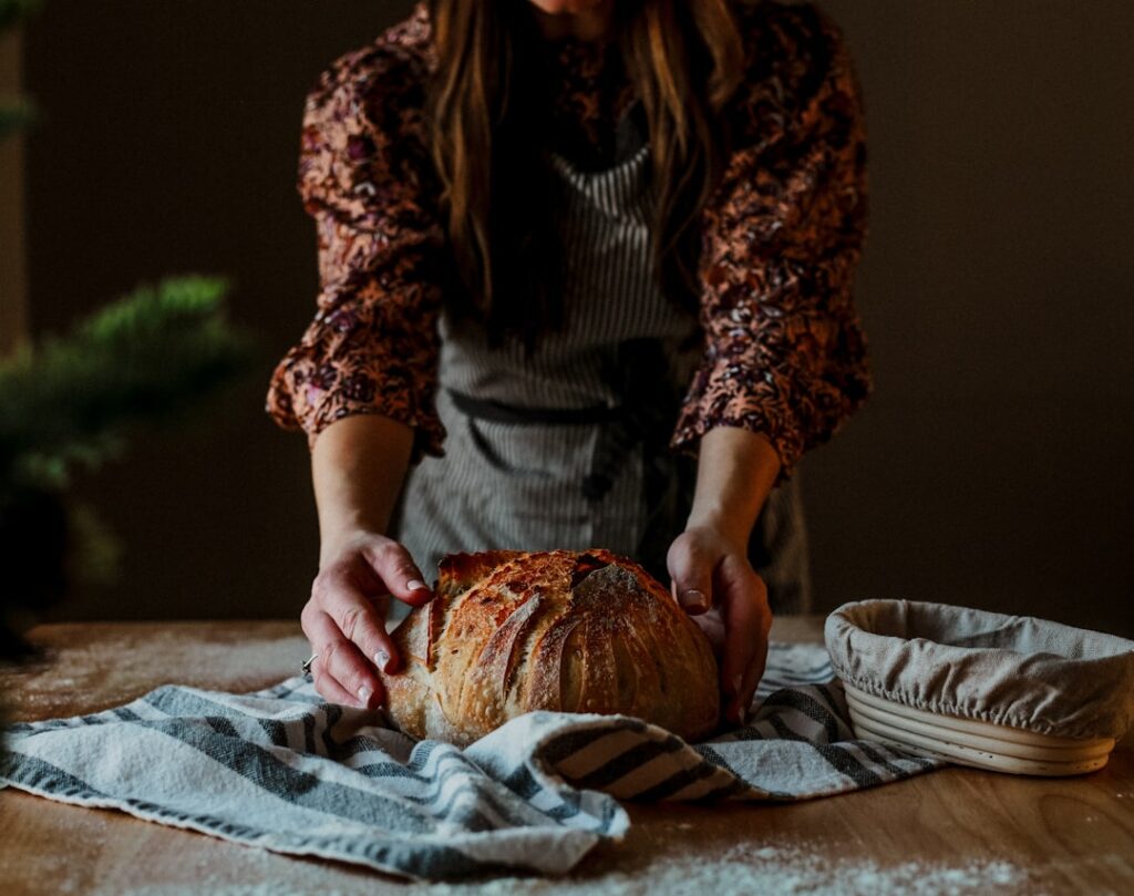 Person in a floral blouse shapes a round rustic loaf of bread on a striped towel on a wooden table