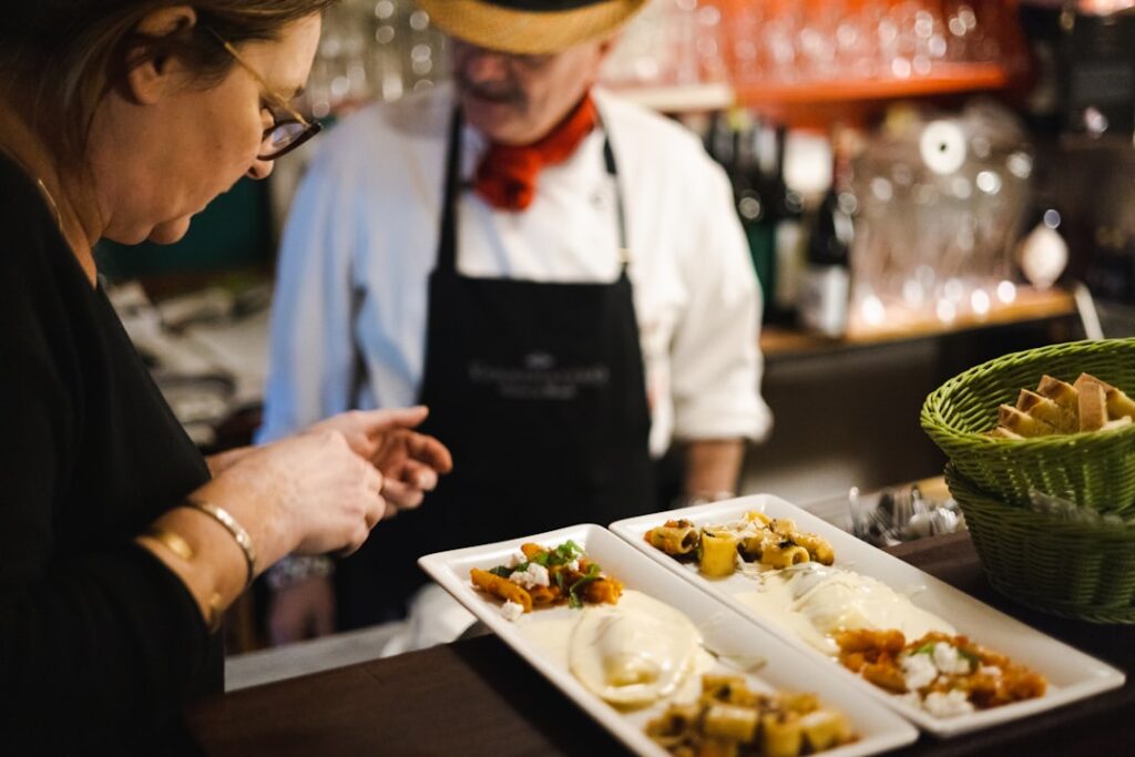 Two staff members in a kitchen plate upscale dishes at a counter, with a green bread basket nearby and plated courses in the foreground