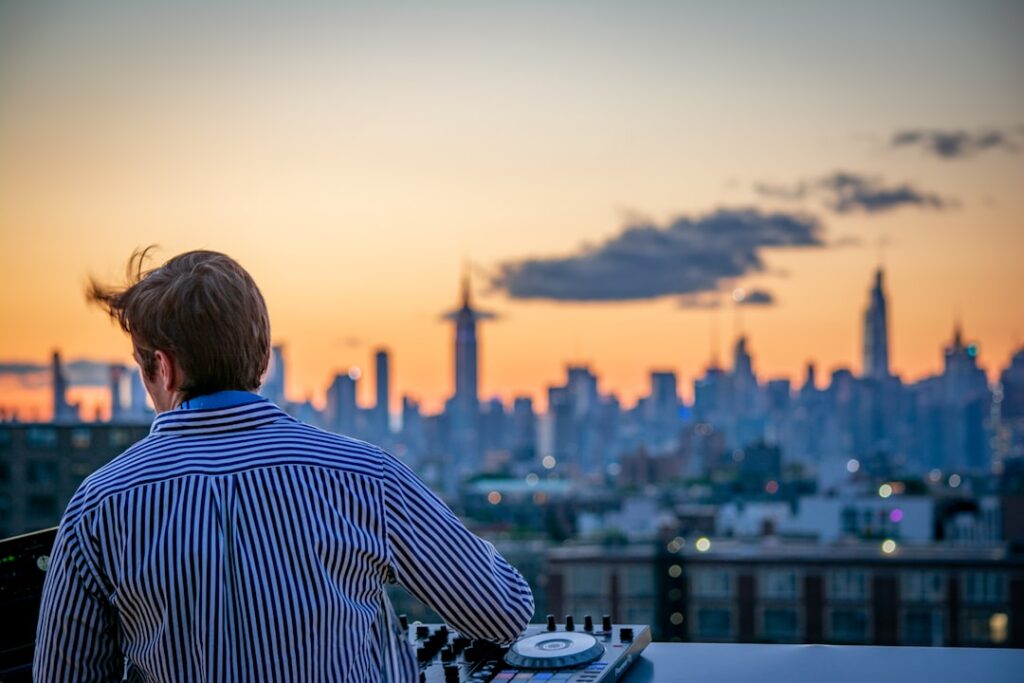 DJ on a rooftop deck mixing music as the city skyline glows at sunset behind him, wearing a striped shirt.