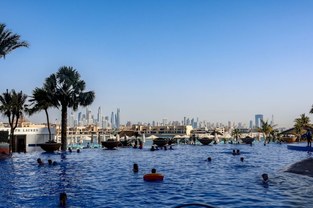People swimming in a large outdoor pool with palm trees and a distant city skyline under a clear blue sky.
