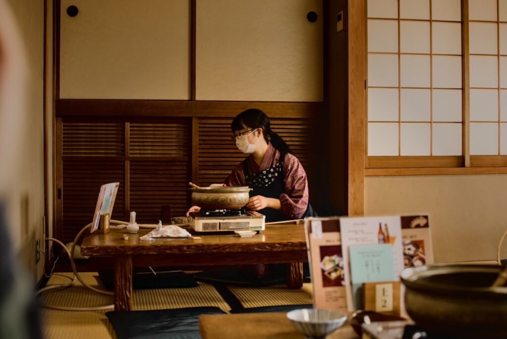 Woman wearing a mask cooks at a low wooden table on a portable stove in a traditional Japanese room with tatami mats and shoji screens.