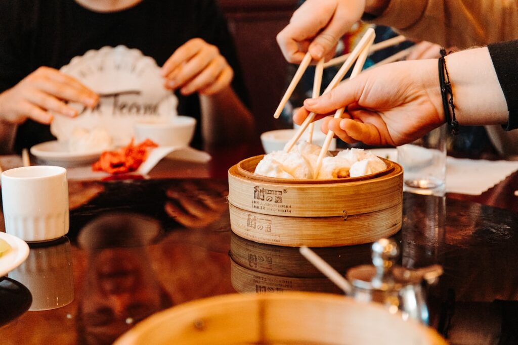 People using chopsticks to pick dumplings from a wooden steam basket at a dim sum meal.
