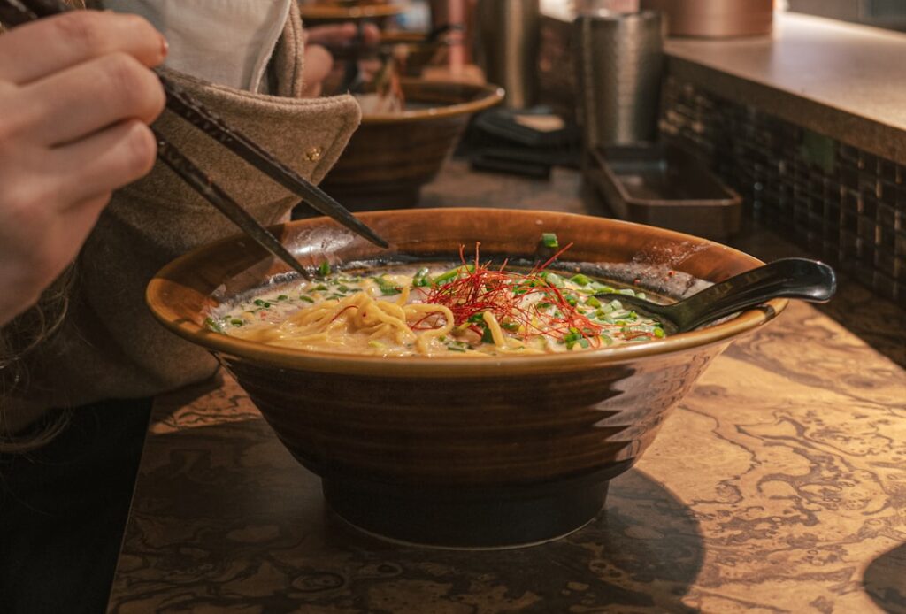 A bowl of ramen on a marble table, greens and red chili threads on top, with chopsticks lifting noodles and a spoon resting in the bowl.