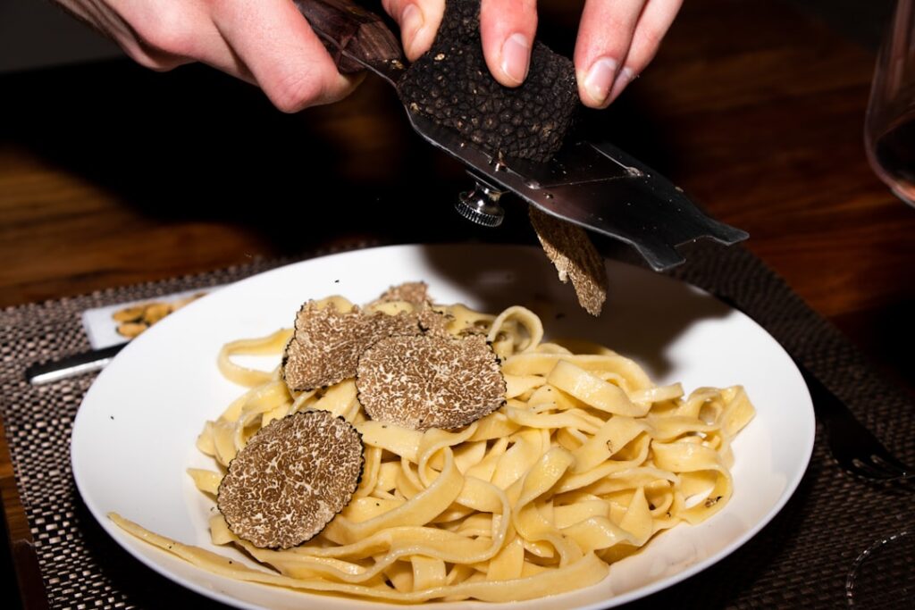 Fettuccine pasta on a white plate with thin truffle shavings being shaved over it by a hand-held slicer.