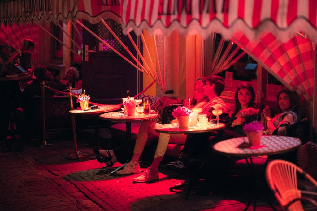 Group of friends seated at round outdoor tables under a striped canopy, drinks and flowers on the tables in pinkish evening light.
