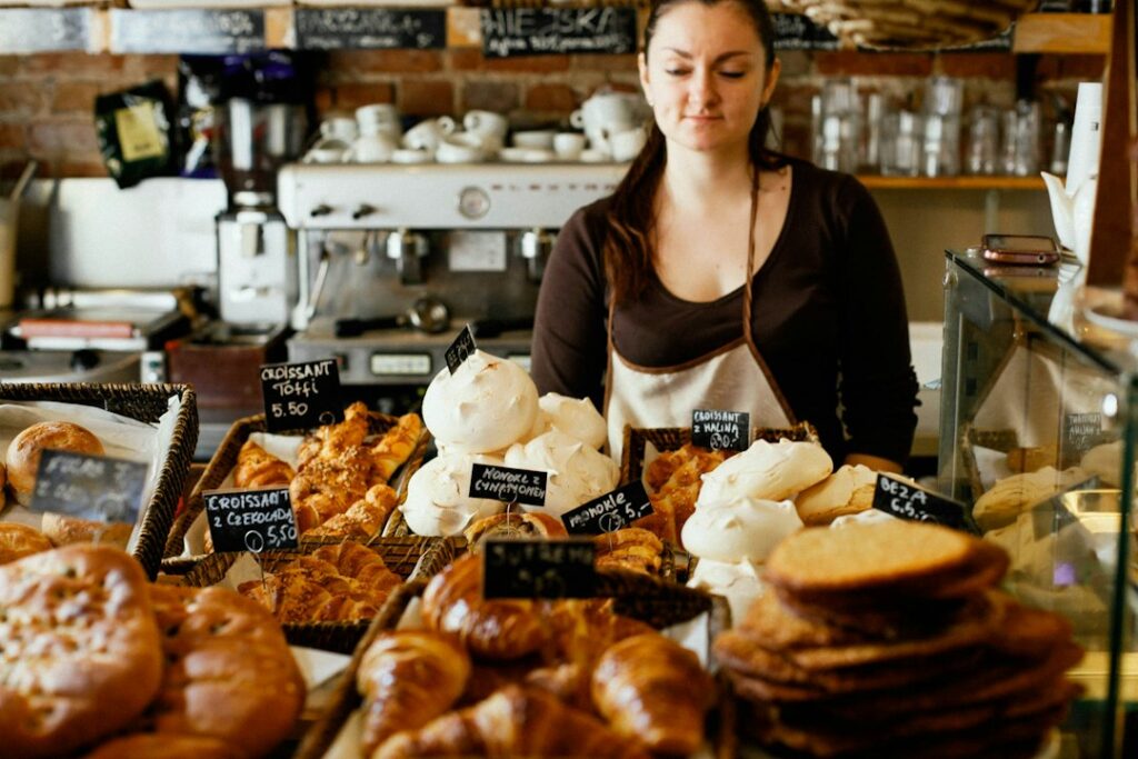 Female bakery worker in a brown shirt and apron stands behind a glass display filled with croissants and pastries.