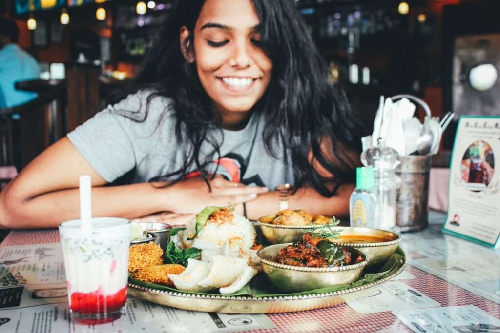 Young woman smiles while seated at a table with a large thali-style Indian meal and a glass of beverage in a restaurant setting.