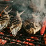 Two whole grilled fish with charred skin on a barbecue grill, with skewered red sausages and meat chunks beside them.