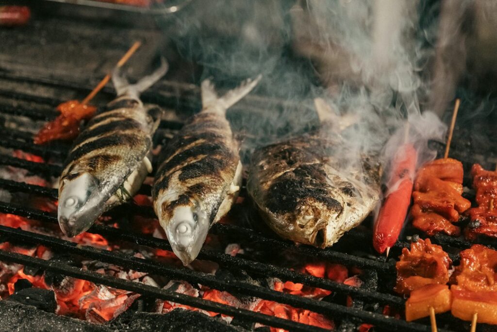 Two whole grilled fish with charred skin on a barbecue grill, with skewered red sausages and meat chunks beside them.