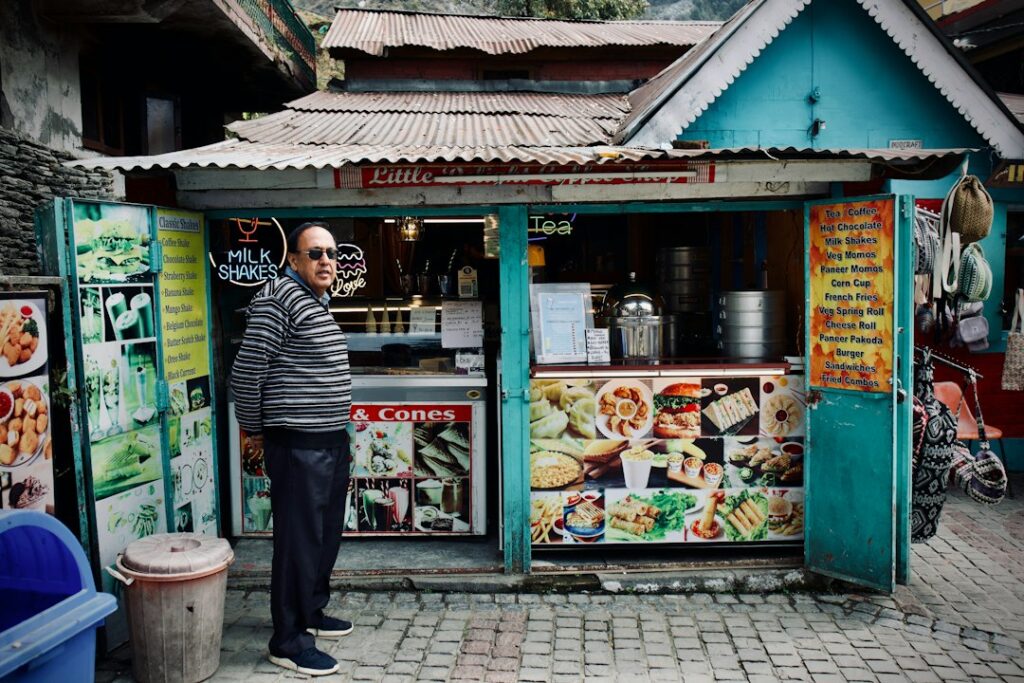 Man in a striped sweater stands at a teal roadside snack stand offering milkshakes and cones, facing the camera.